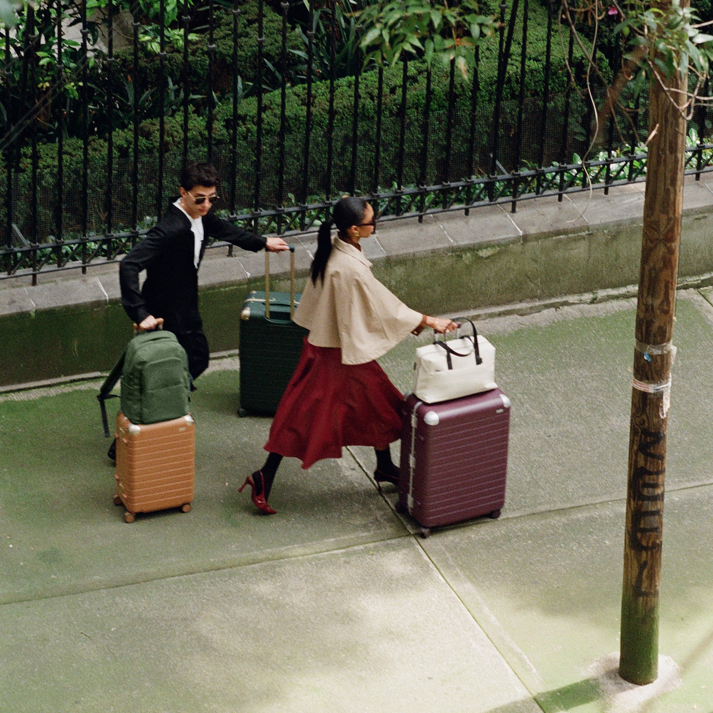 Two people pulling suitcases on a sidewalk with a fence and tree in the background.