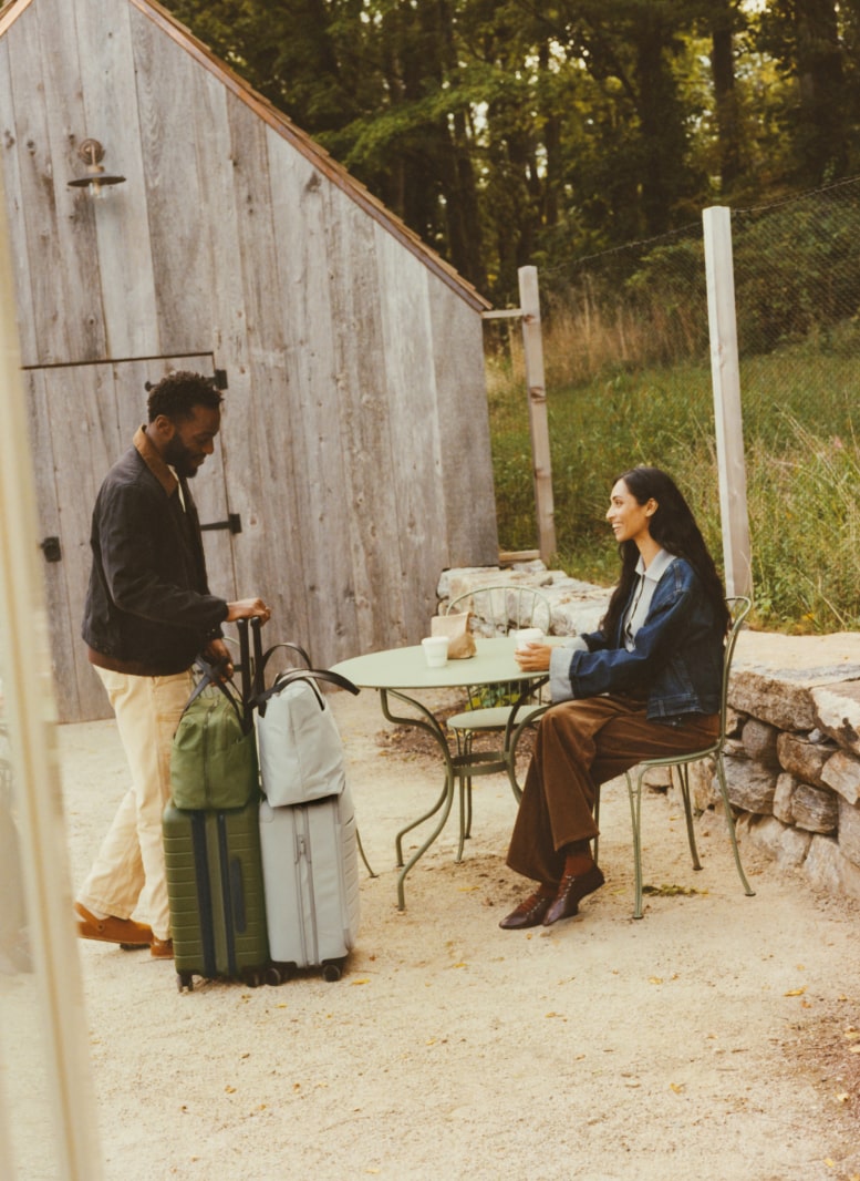 Two people sitting outside of a vacation cabin with two sets of Away luggage in green and gray