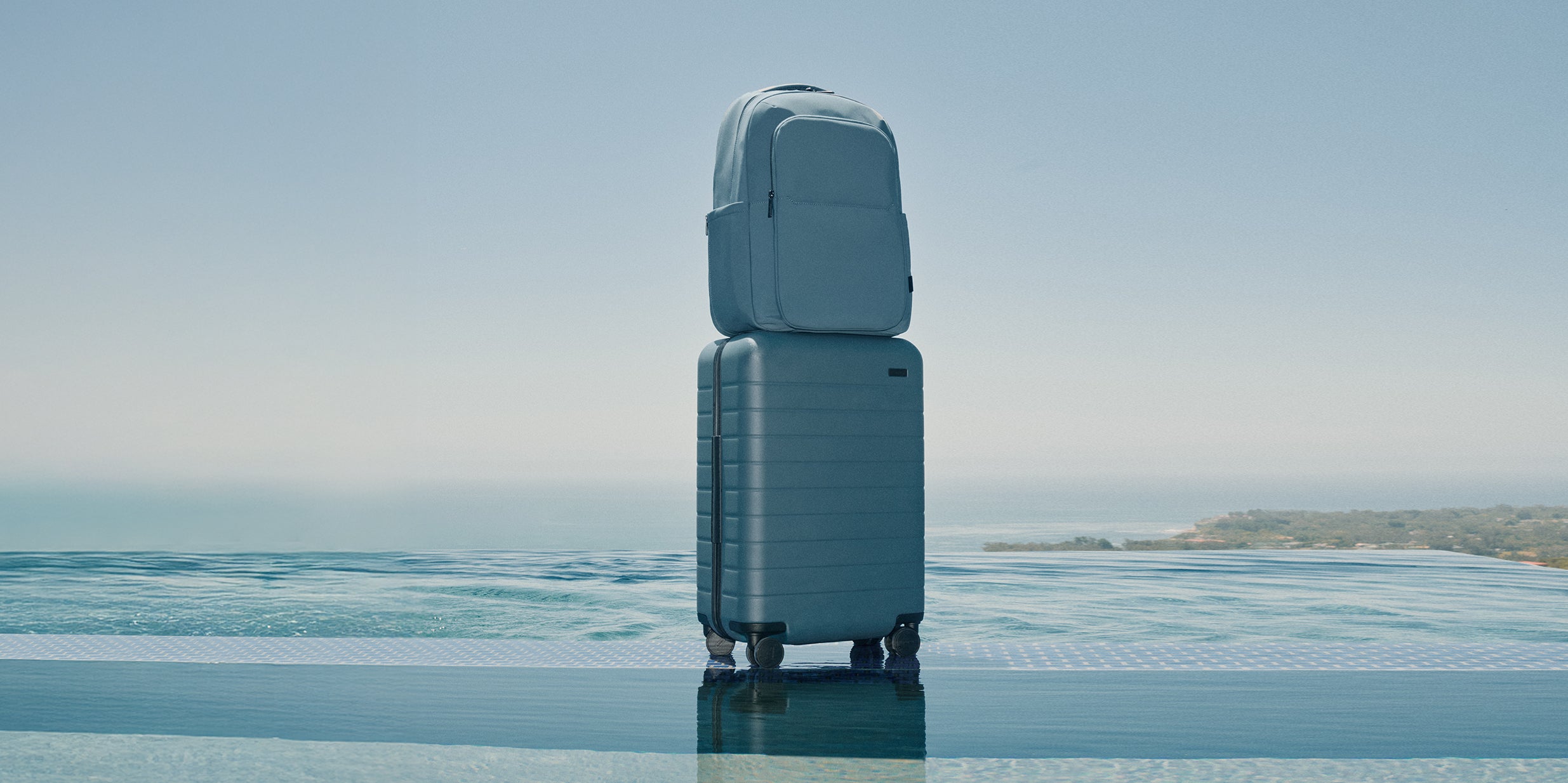A Coast Blue Commuter Backpack stacked on a Coast Blue suitcase with a reflective surface and a blurred landscape in the background