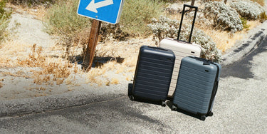 Three hard-shell rolling suitcases standing on a paved road outdoors, with a blue directional sign and dry vegetation in the background. The suitcases appear to be the product being shown in a real environment.