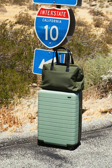 A mint green hardshell suitcase standing on a road with a dark olive green duffel bag resting on top, positioned in front of Interstate 10 California road signs and desert vegetation.