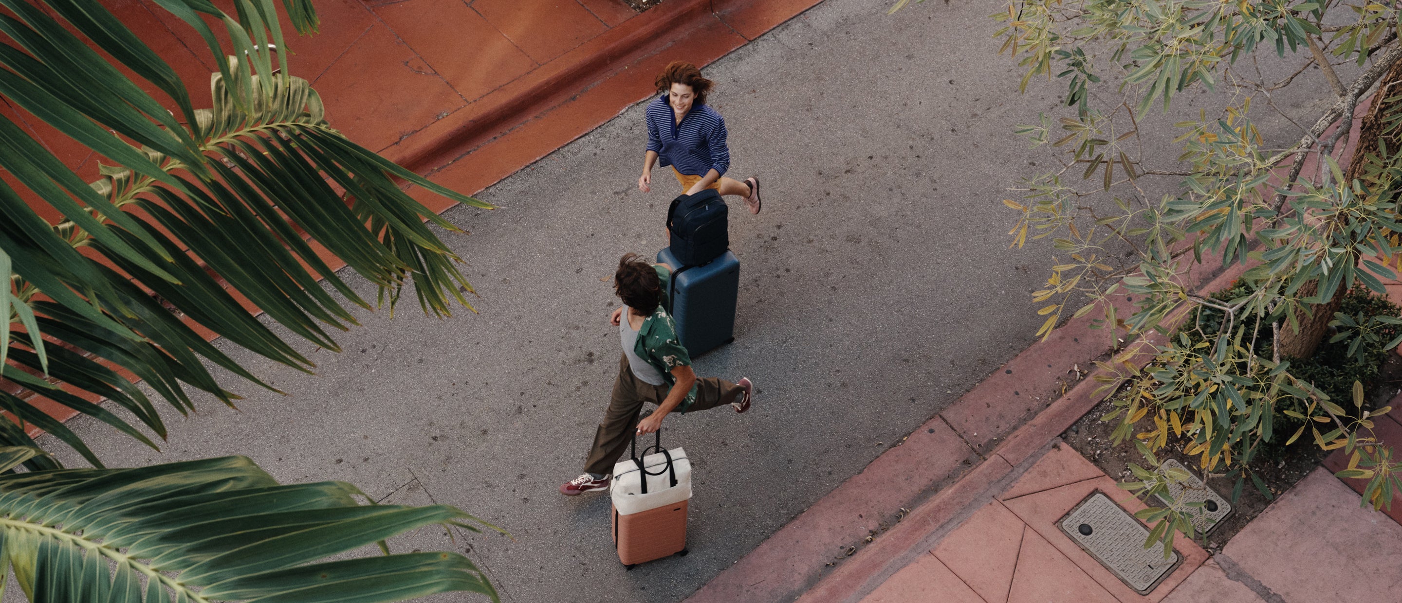 Male model running with The Carry-On in Clay Pink and The Everywhere Bag in Salt White next to female model running with The Commuter Backpack in Navy Blue stacked on The Trunk in Coast Blue
