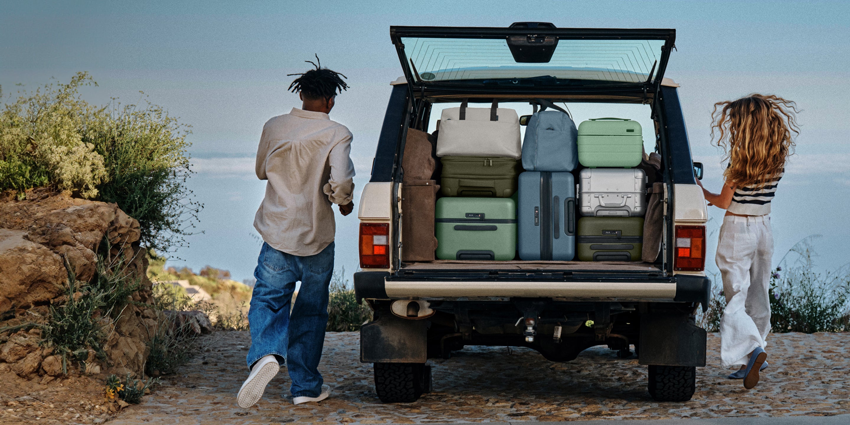 Two people standing next to a car with open trunk full of suitcases by a scenic beach.
