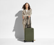 A 5'6" female model is standing next to The Bigger Carry-On suitcase and leaning her arms on the raised handle to show size and scale
