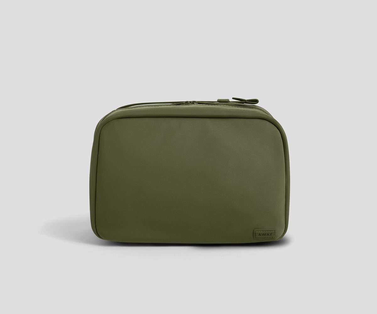 An olive green zippered toiletry bag with a small dark brand tag on the bottom right corner, photographed on a neutral light gray background.
