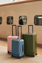 Three suitcases in pink, blue, and green in front of a beige train.
