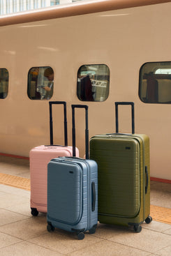 Three suitcases in pink, blue, and green in front of a beige train.