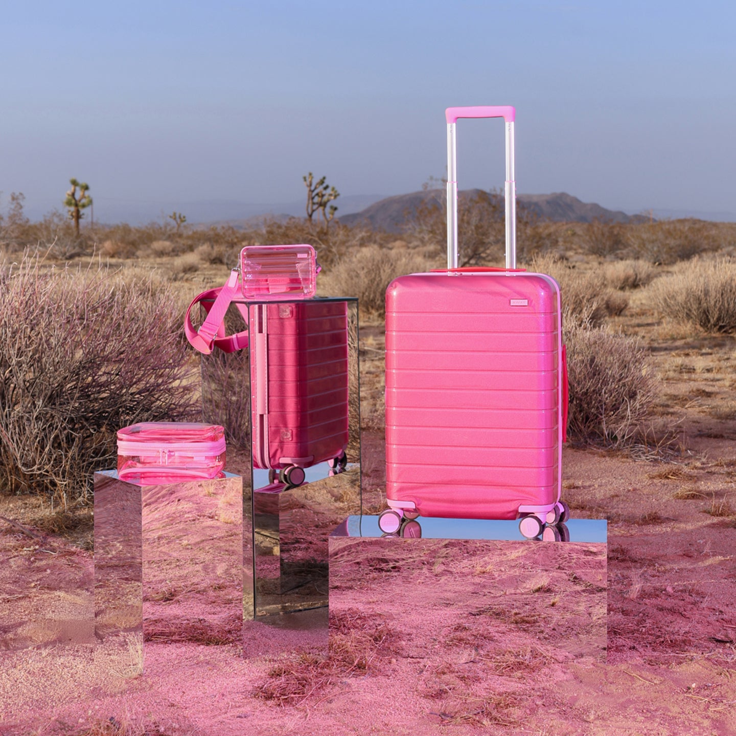 Front product view of The Bigger Carry-On in Translucent Pink, The Mini Crossbody in Translucent Pink and Clear Toiletry Bag in Translucent Pink shot in the desert