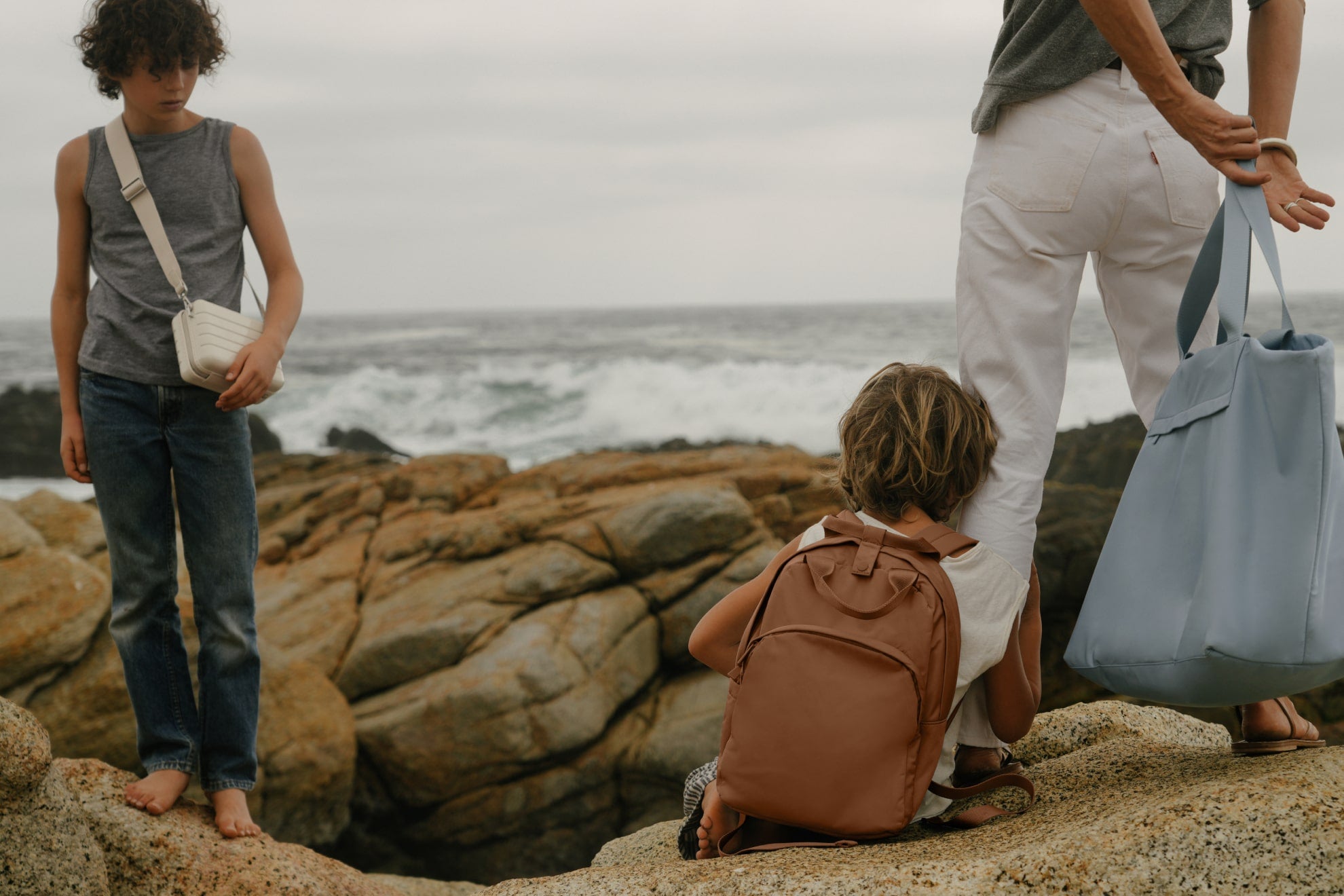 Family of three on a rocky beach with The Mini Crossbody in Salt White, The Featherlight Backpack in Amaro Brown, The Large Featherlight Tote in Arctic Blue