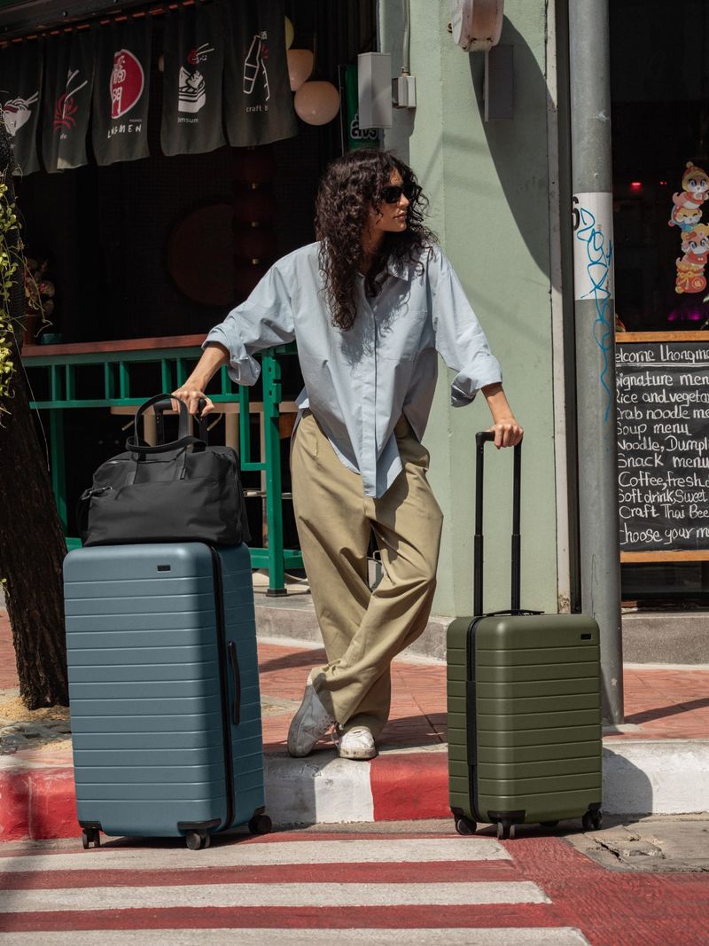 A woman in the streets of Bangkok with The Trunk in Coast Blue, The Carry-On in Olive Green, The Everywhere Bag in Black